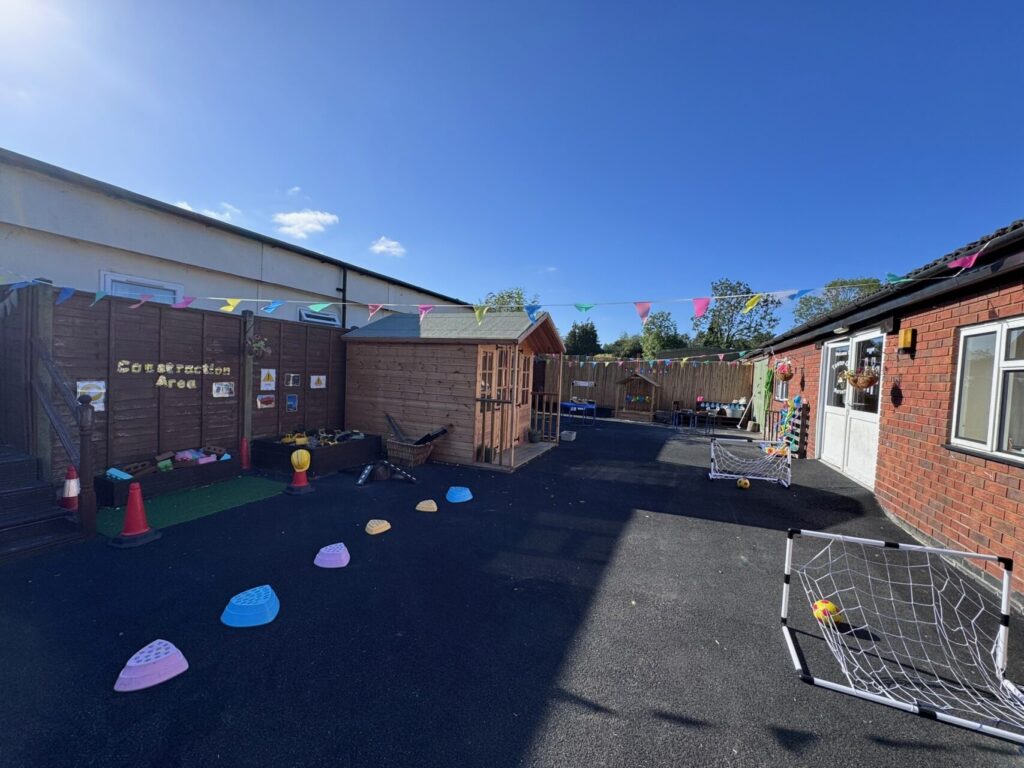 Outdoor play area with colorful bunting, small soccer nets, stepping stones, and a wooden construction zone. Sunny day with a cheerful, inviting vibe.