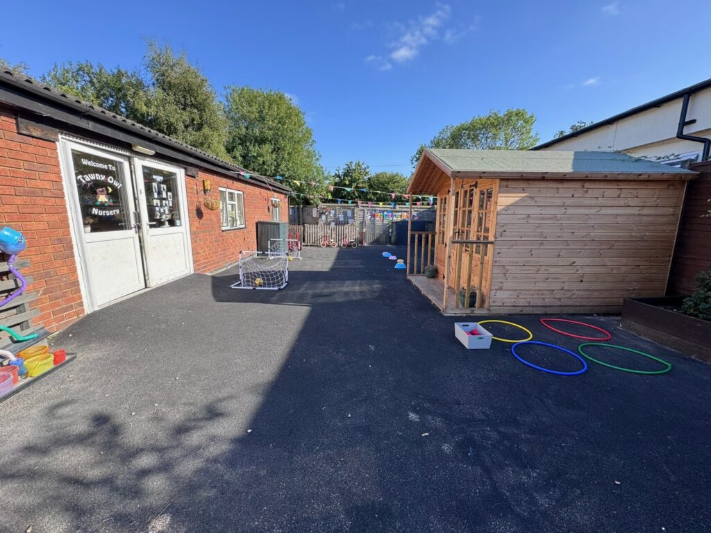 Outdoor nursery playground with colorful hoops on blacktop, wooden playhouse on the right, brick building on the left. Clear sky, sunny and inviting.