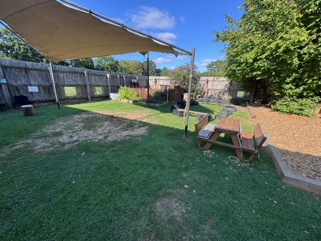 Outdoor play area with fake grass, shaded by a white canopy. Features toy tools, a small bench, a pallet table, and cushions, surrounded by trees.