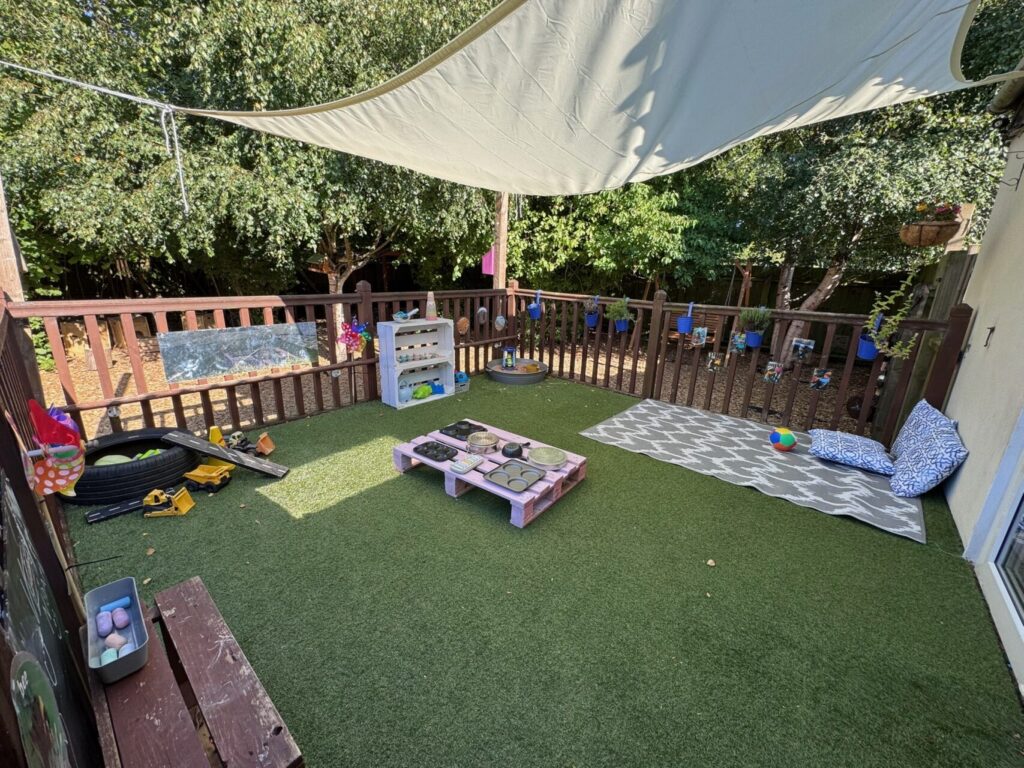 Outdoor play area with fake grass, shaded by a white canopy. Features toy tools, a small bench, a pallet table, and cushions, surrounded by trees.