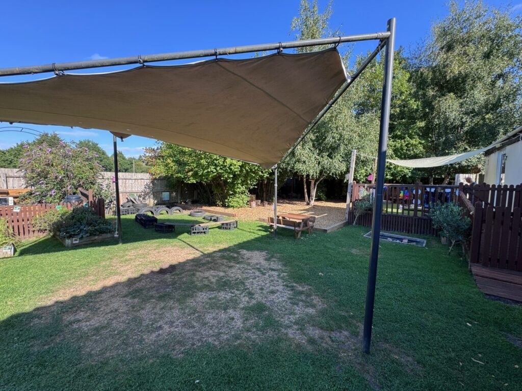 A sunny backyard with green grass, large canopy shades, and seating made from wooden pallets. Trees and a wooden fence line the peaceful area.