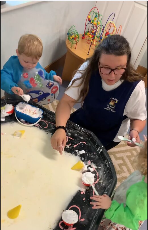 A woman and two children engage in a playful sensory activity with white foam and colorful items on a black tray. The setting feels joyful and educational.