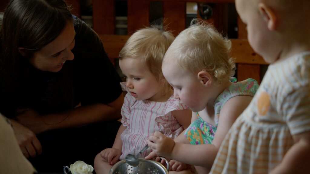 A woman smiles at three toddlers focused on a toy, creating a warm, nurturing scene. The children wear colorful outfits, suggesting a playful atmosphere.