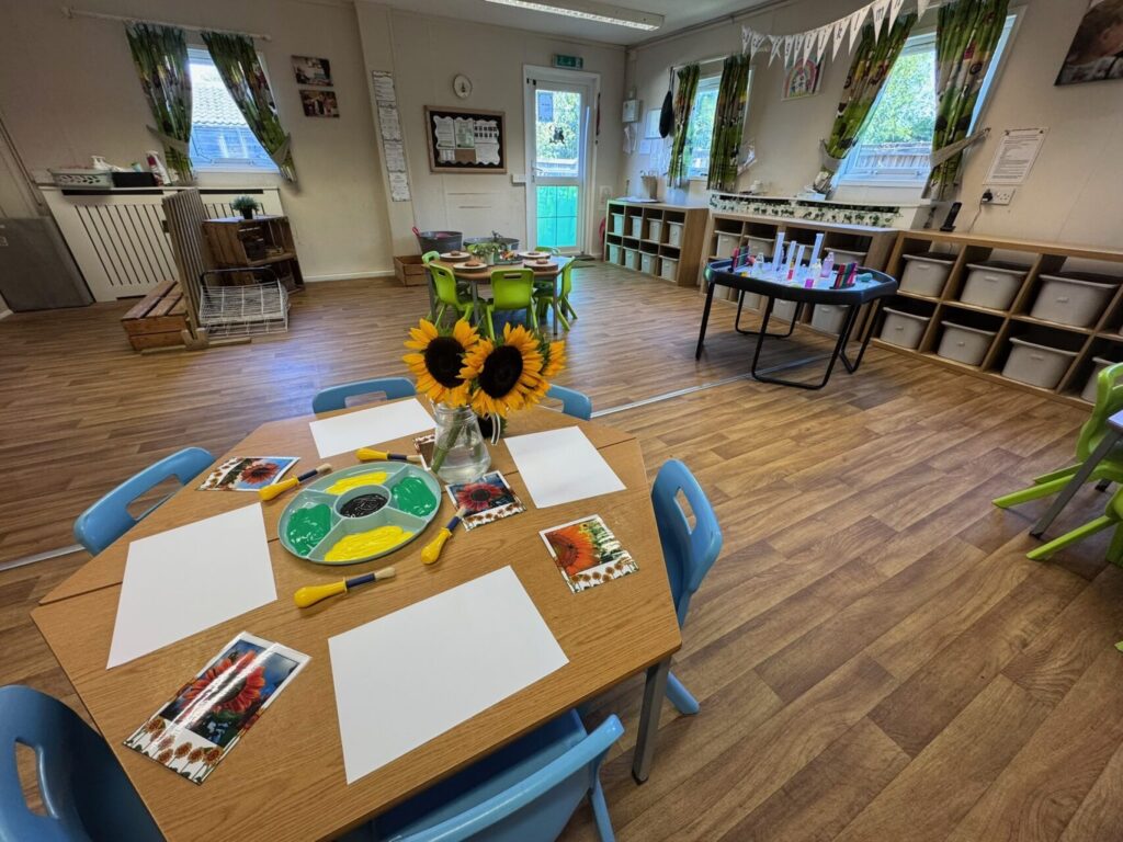 Classroom with wooden floors, art supplies on tables surrounded by blue chairs. Sunflowers in a vase add brightness. Shelves with bins line the walls.