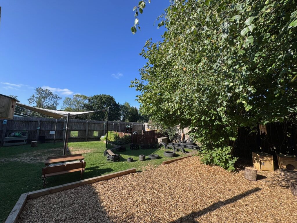 Sunny outdoor play area with green grass, wooden benches, and tires under a large tree. Wood chips cover part of the ground. Peaceful ambiance.