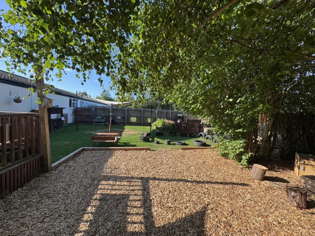 Sunny outdoor play area with wood chips, shaded by trees. Includes benches, a lawn with play equipment, and a quiet, inviting atmosphere.