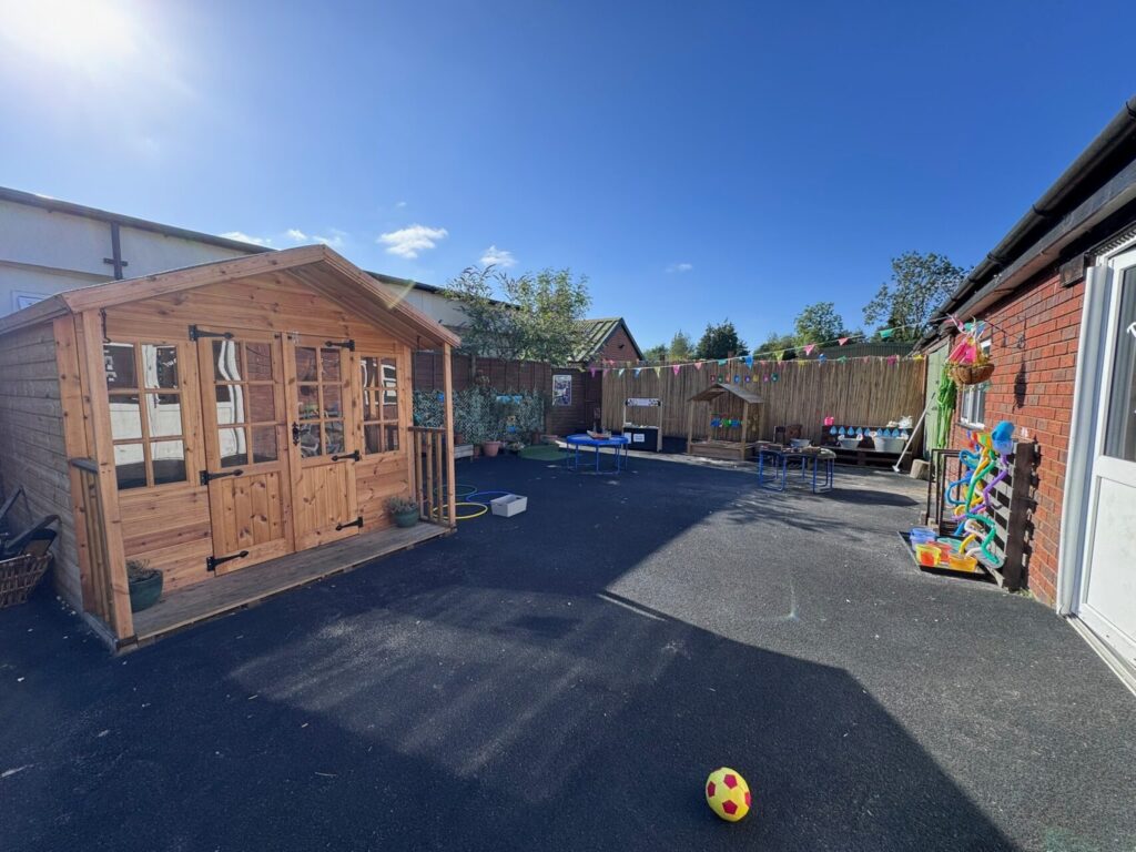 A sunny outdoor play area with a wooden playhouse, colorful toys, and tables. Festive bunting hangs on the fence, evoking a cheerful, welcoming atmosphere.