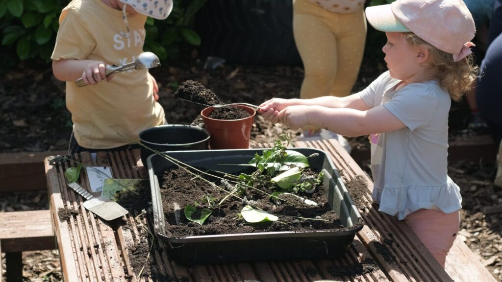 A person gestures while talking to children in a garden. One child wears a red cap, another a sun hat. The scene is lively and educational.