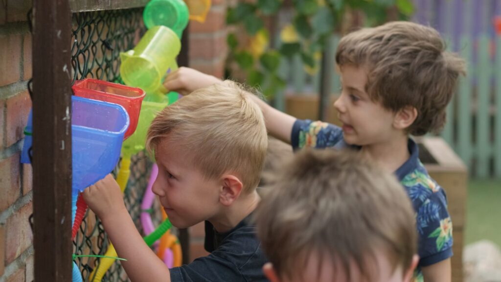 A person gestures while talking to children in a garden. One child wears a red cap, another a sun hat. The scene is lively and educational.