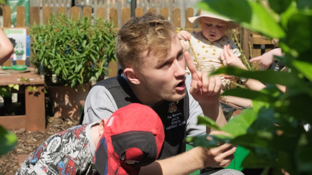 A young man interacts with children in a garden, gesturing and talking animatedly. A child in a red cap and another in a sunhat are engaged nearby.