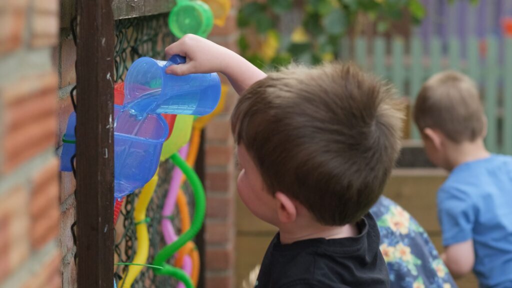 A young boy pours water from a blue cup into a colorful outdoor water play structure, concentrating intently. Another child is in the background playing.