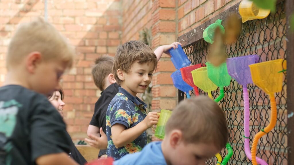 Children playing with colorful funnels on a chain-link fence. They appear engaged and happy, wearing casual clothes, outside on a sunny day.